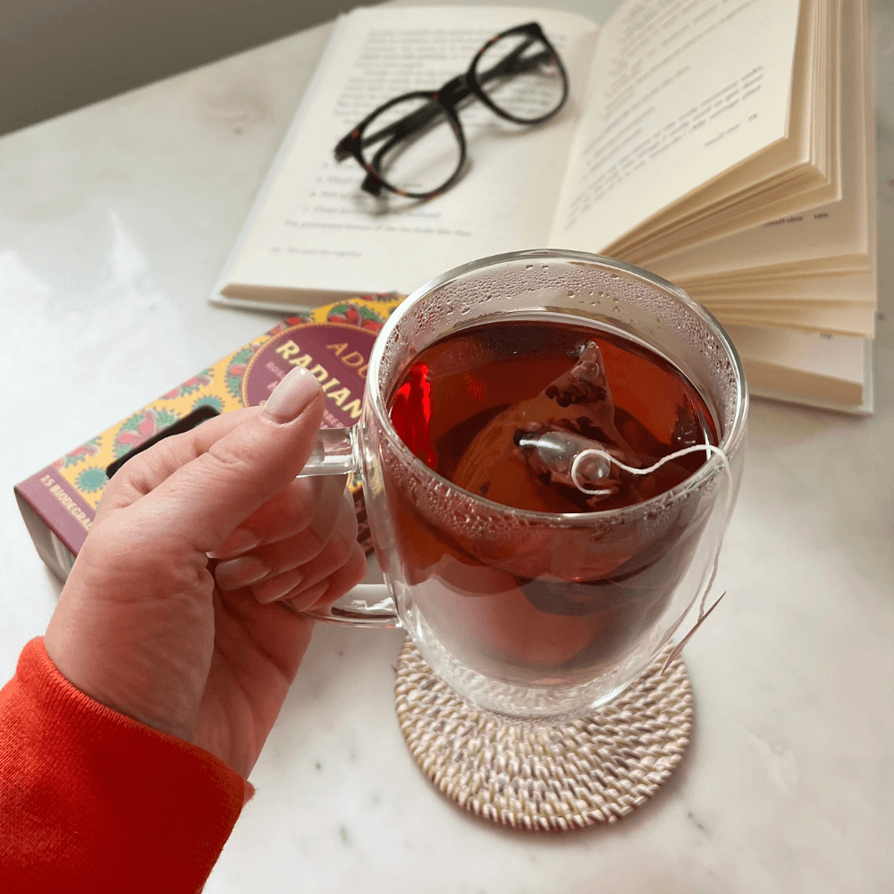 Hand holding a glass mug of Aduna Radiance tea with a book and glasses in the background