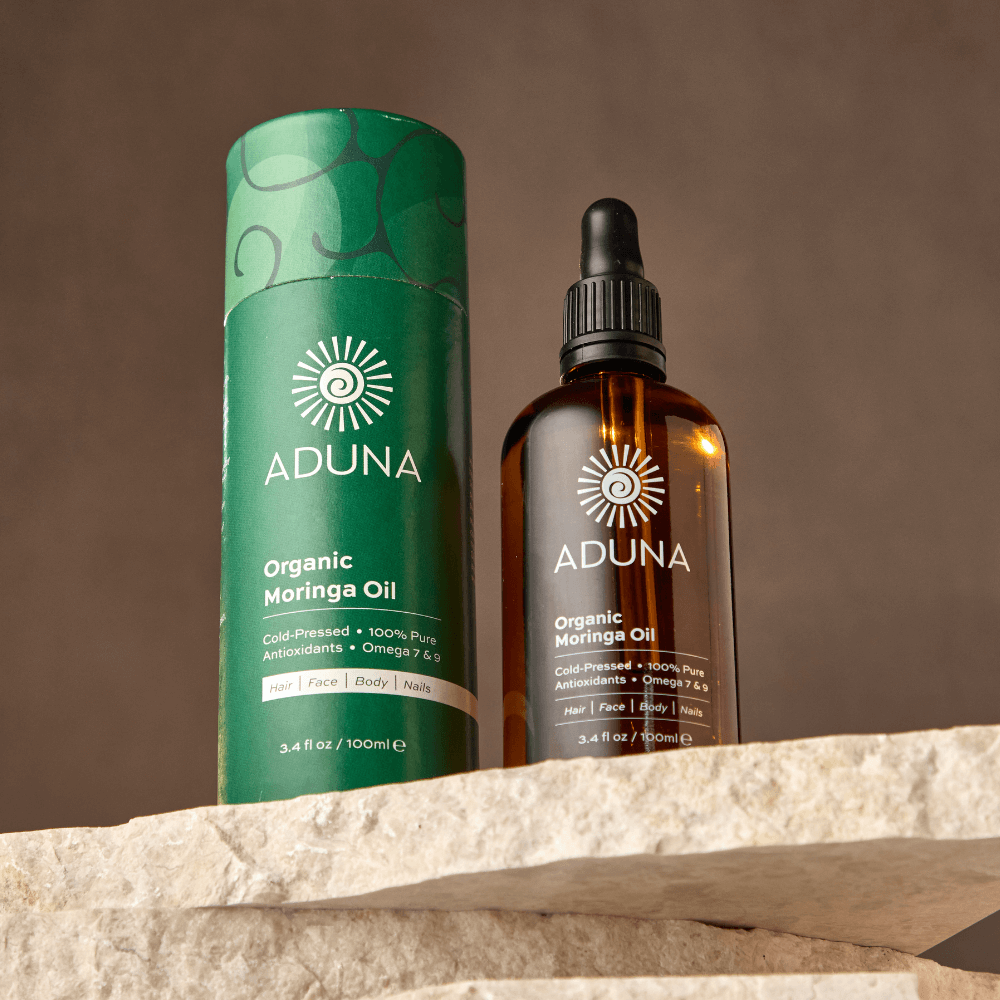 Aduna Organic Moringa Oil bottle and green cylindrical container on a stone surface with a brown background
