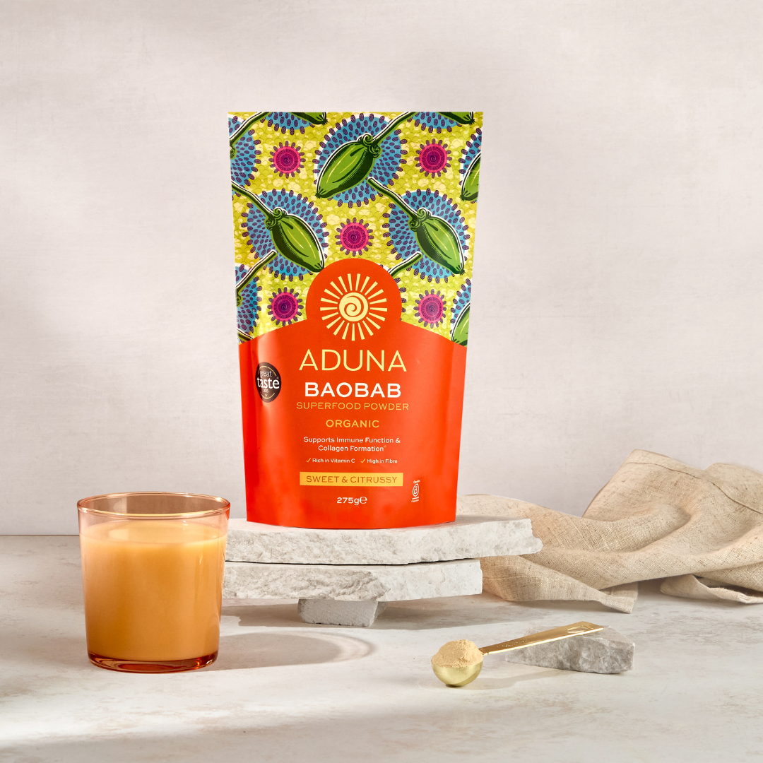 Aduna Baobab Pouch displayed on stacked marble stands, next to an orange drink and a spoon of baobab powder on a marble surface