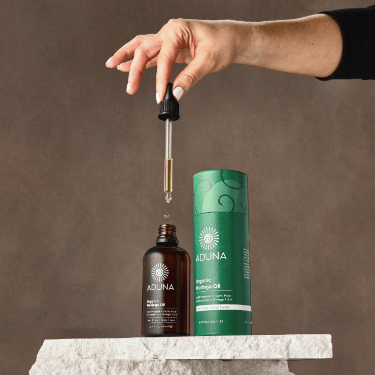 Hand using a dropper to extract Aduna Organic Moringa Oil from a bottle on a marble surface with a brown background.