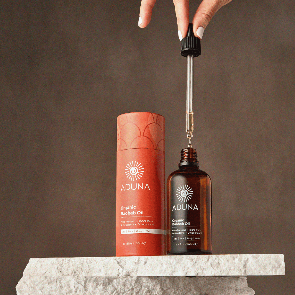 Aduna Organic Baobab Oil bottle with dropper and packaging on a stone surface against a brown background