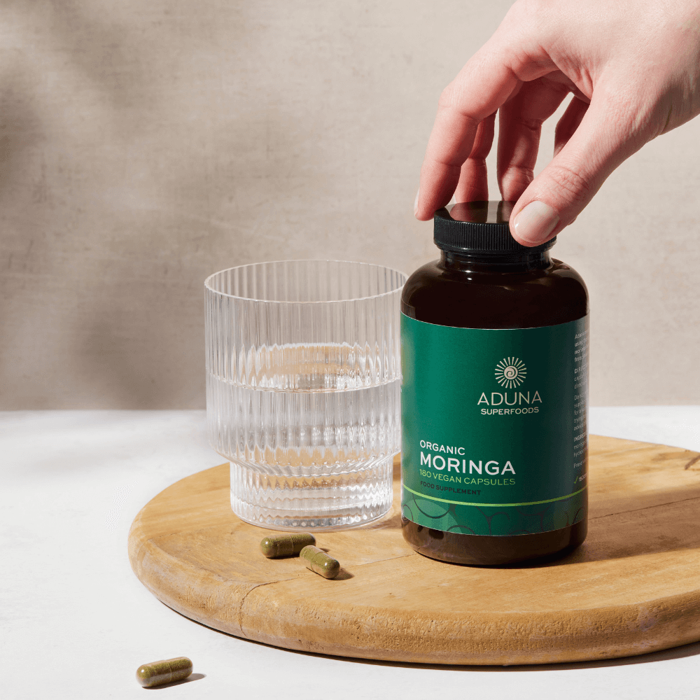 Hand opening a bottle of Aduna Superfoods Organic Moringa capsules on a wooden board with a glass and three capsules laying next to it.