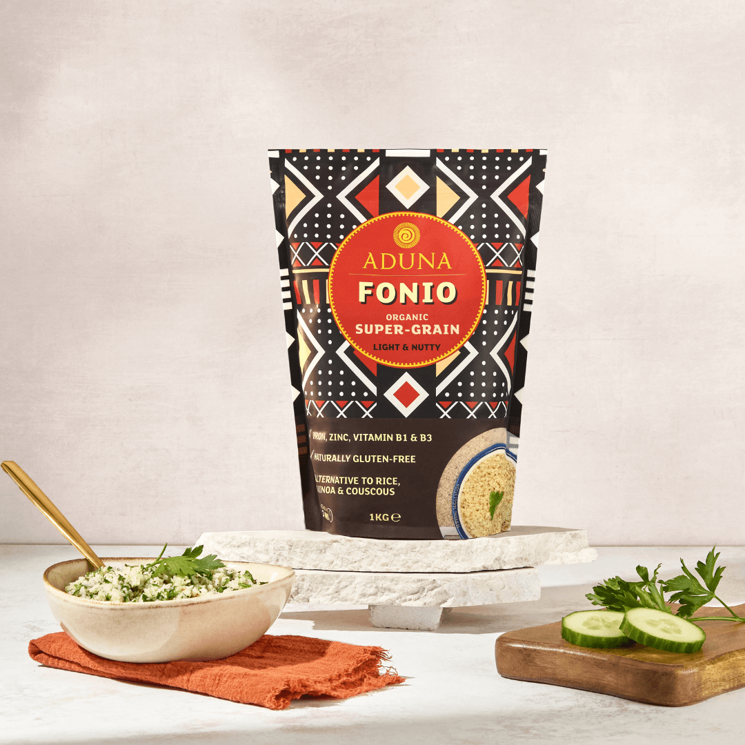 Aduna Fonio Grain 1kg Pouch displayed on marble stands. A bowl of a cooked fonio salad and a chopping board with cucumbers in the front.