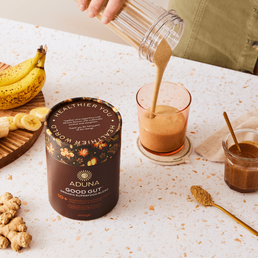 A package of Aduna Good Gut Organic Superfood Blend on a kitchen bench next to someone pouring a blend into a glass