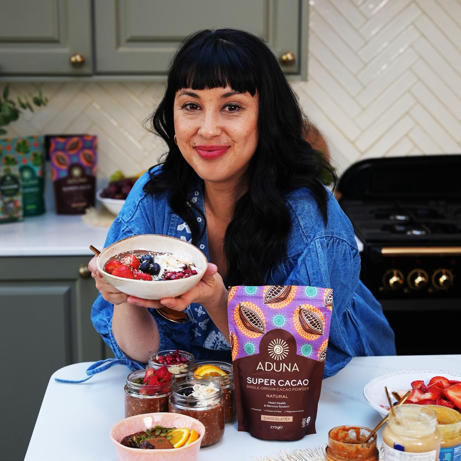 Melissa Hemsley holding a bowl of fruit and a package of Aduna Super Cacao in a kitchen.