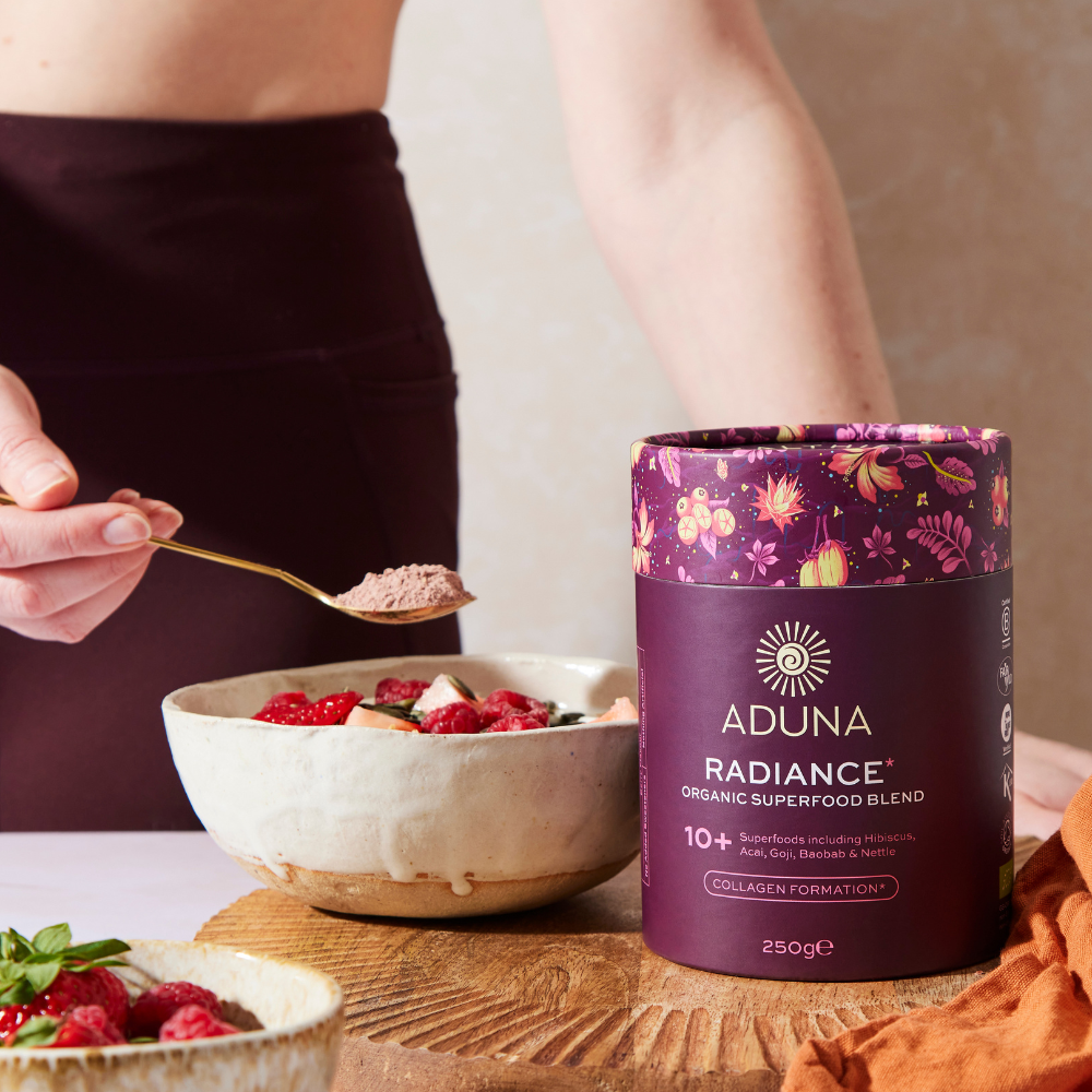 Woman holding spoonful of Aduna Radiance Organic Superfood Blend over a bowl of fruit, with a 250g package of the powder blend next to it