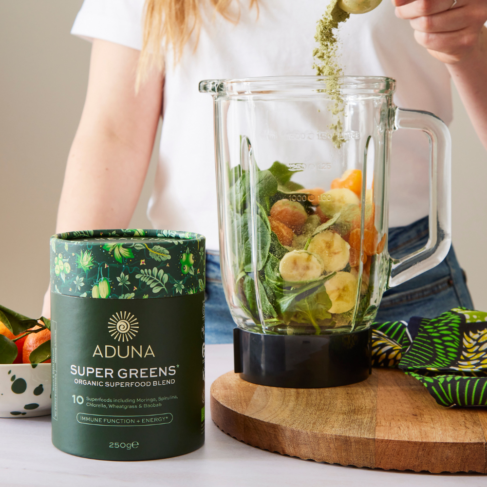 Woman preparing a green smoothie with Aduna Super Greens blend in a blender.