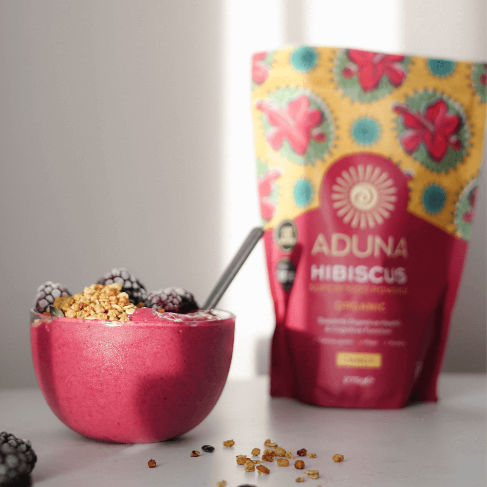 Aduna Hibiscus Superfood Powder 275g package stood next to a bowl of pink yoghurt containing fruit and muesli.