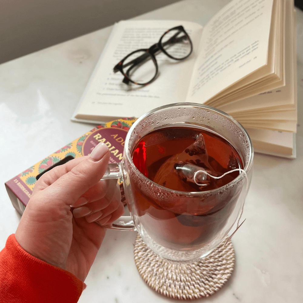 Person holding glass mug of Aduna Radiance Hibiscus Tea with tea bag still inside it