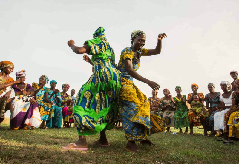 Two African women in colourful traditional attire dancing in a field with onlookers.