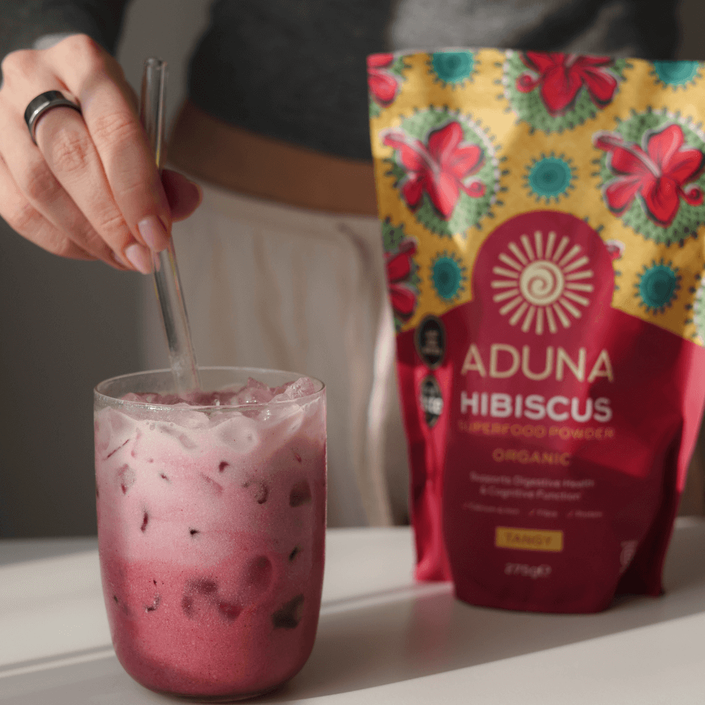 Woman stirring a pink drink with a bag of Aduna Hibiscus powder in the background.