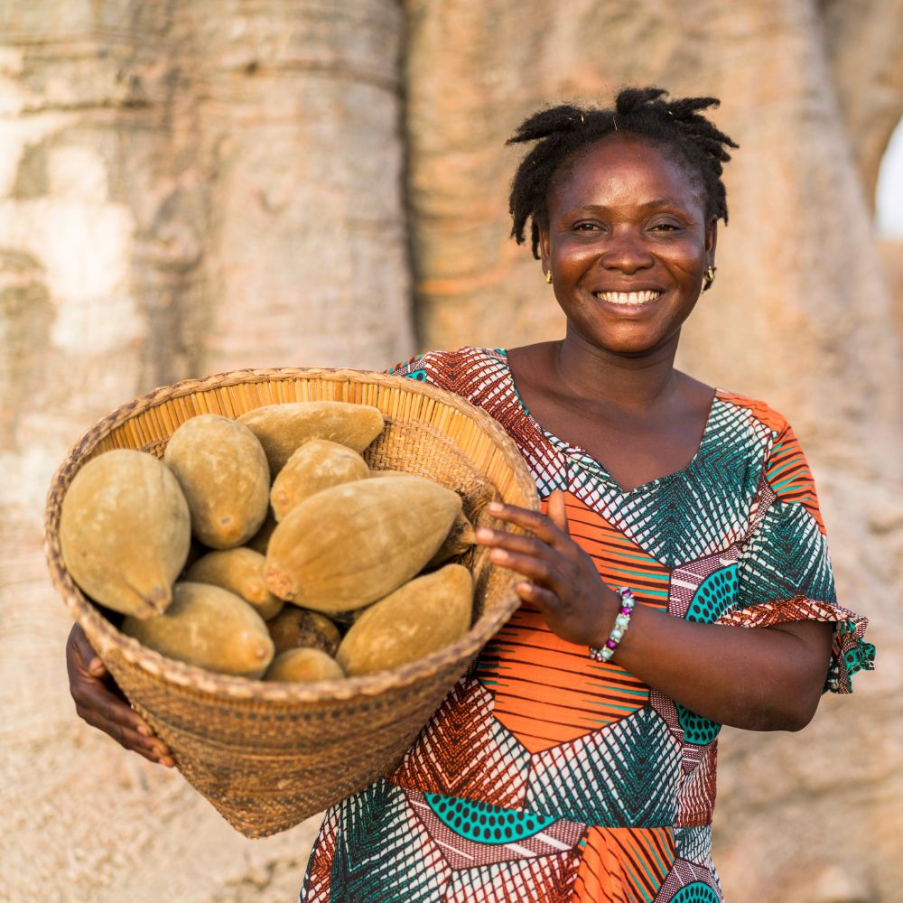 African woman holding a basket of baobab fruits in a natural setting.