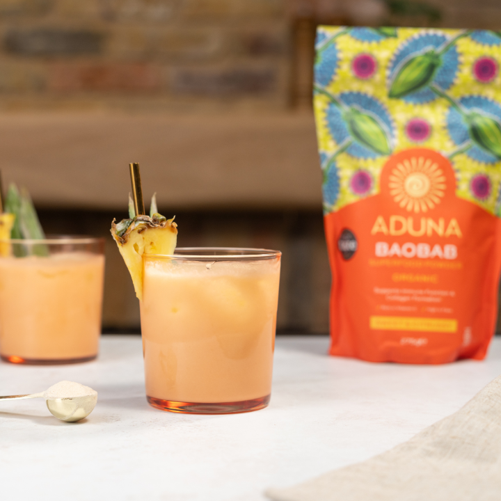 Peach-coloured smoothie with pineapple garnish next to a package of Aduna Baobab Powder on a white surface.