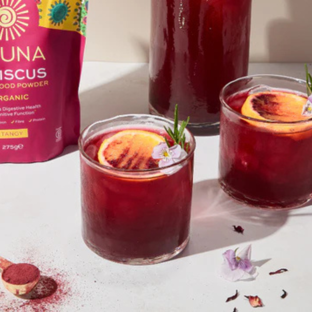 Aduna Hibiscus Organic Superfood Powder 275g package next to a couple of glasses of red juice with garnishes.