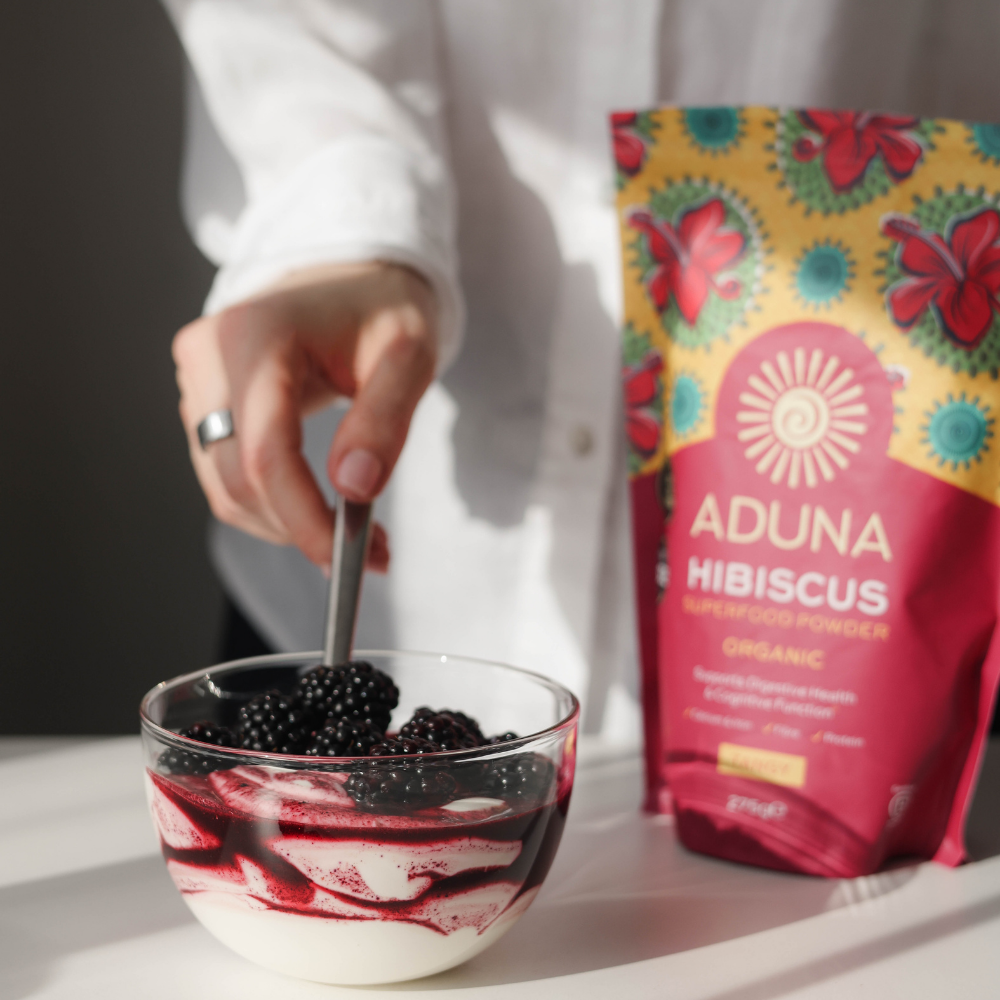 Person stirring a bowl of blackberries with a package of Aduna Hibiscus Superfood Powder in the background.
