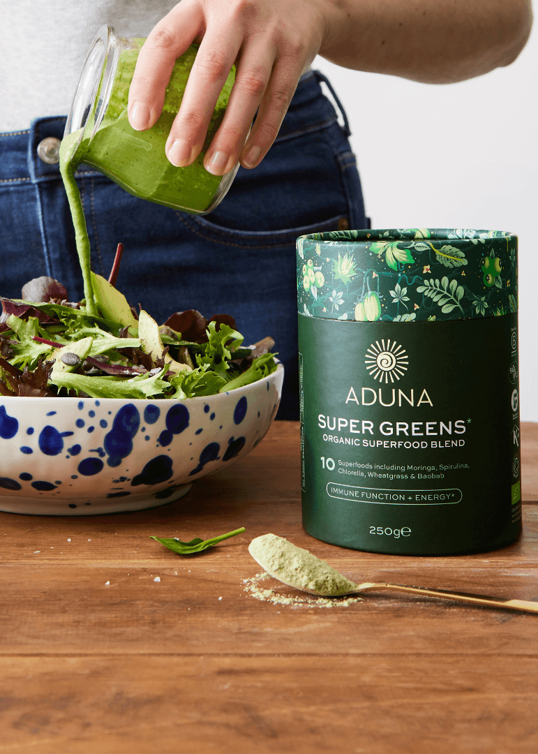 Person pouring Aduna Super Greens superfood into a salad with Aduna Super Greens package on a wooden table.