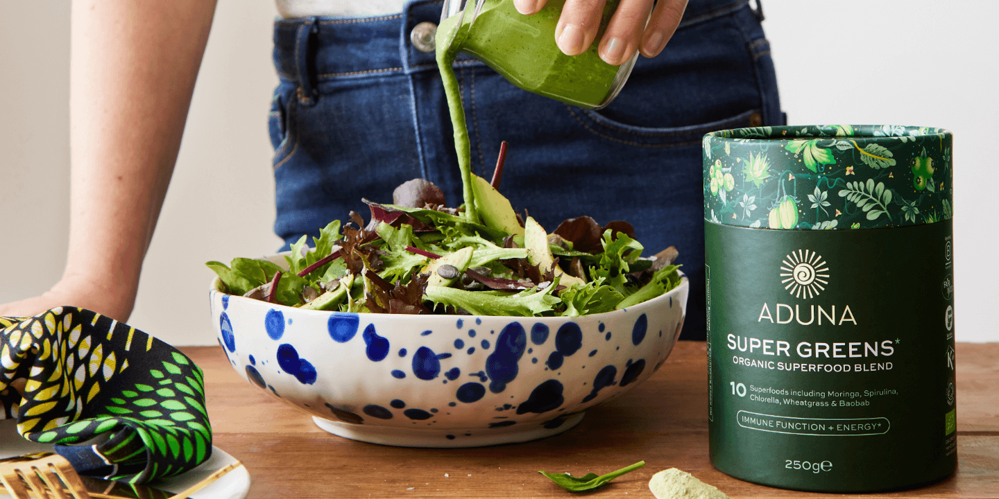 Woman pouring glass of Aduna Super Greens Organic Superfood Blend over a salad as a dressing.