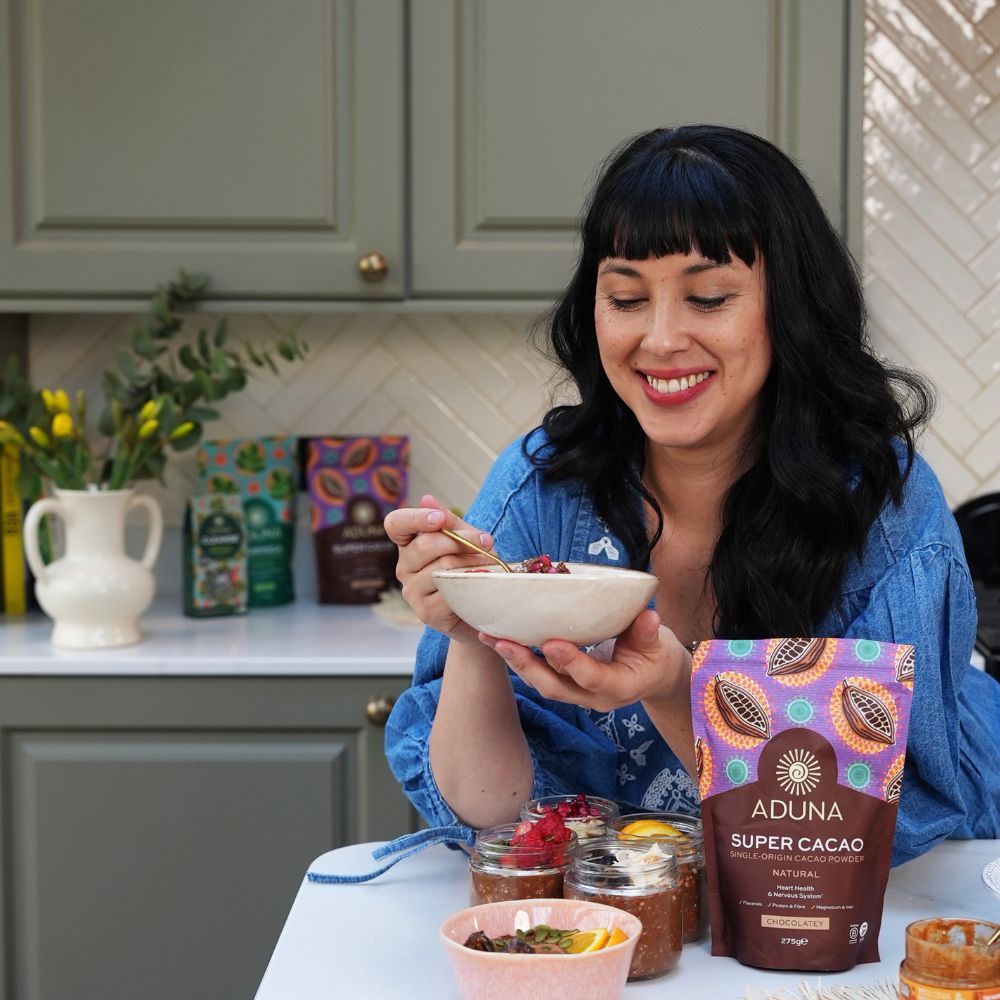 Melissa Hemsley in a kitchen holding a bowl of food with a package of Aduna Super Cacao in front of her.