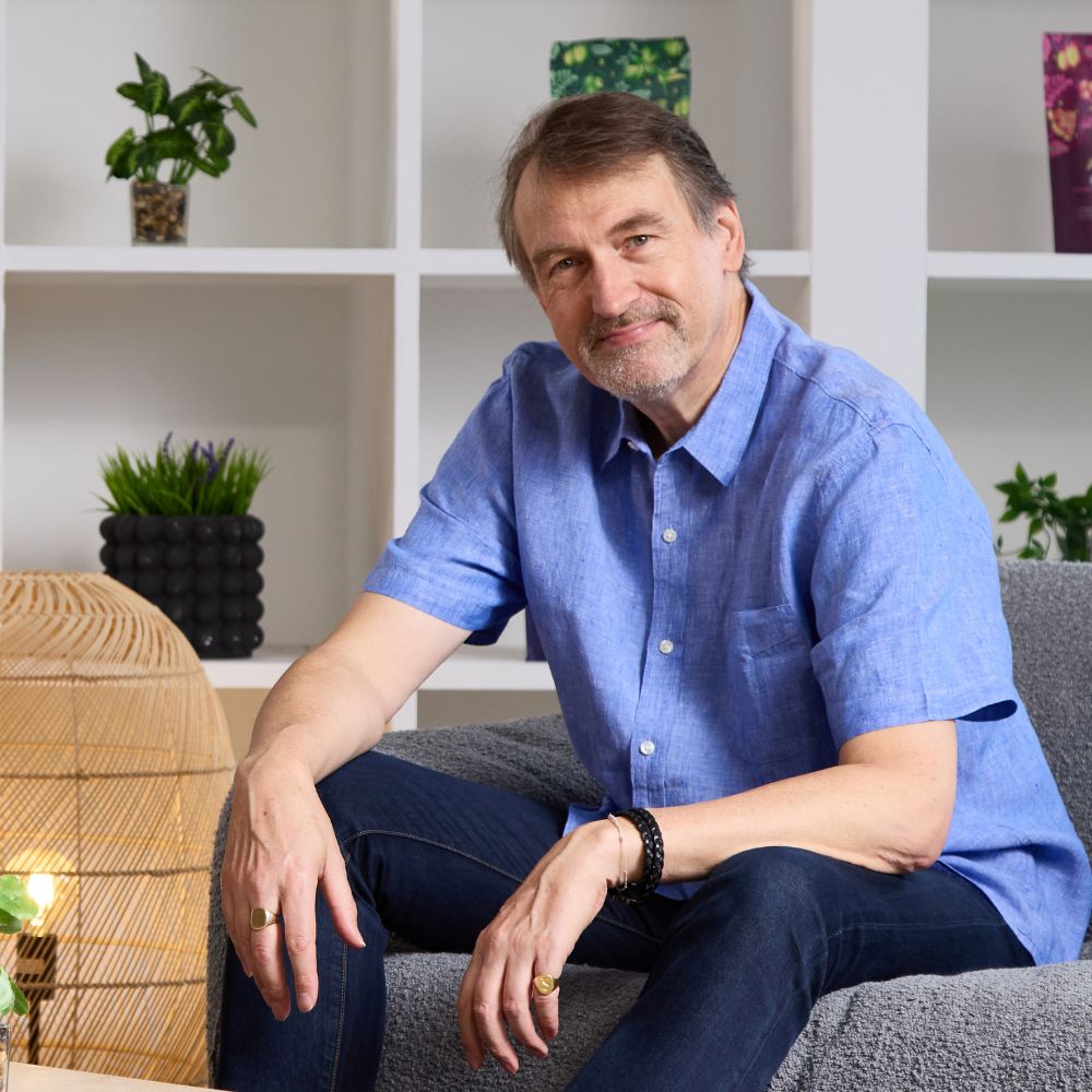 Aduna co-founder sitting on a couch with a white bookshelf and plants in the background.