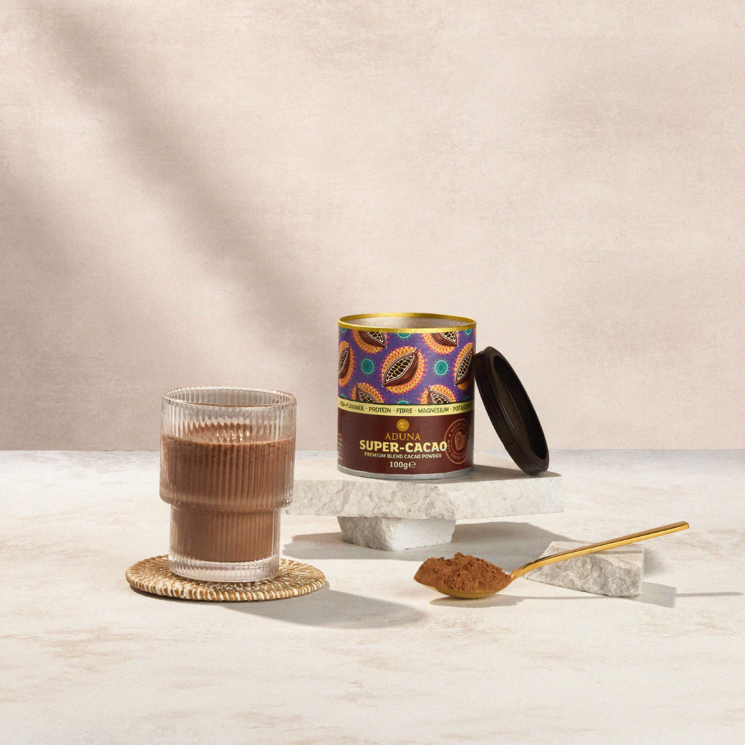 Aduna Super Cacao Powder Tub opened and displayed on a marble stand. In front, a glass of brown chocolate shake and a spoon of brown cacao powder.